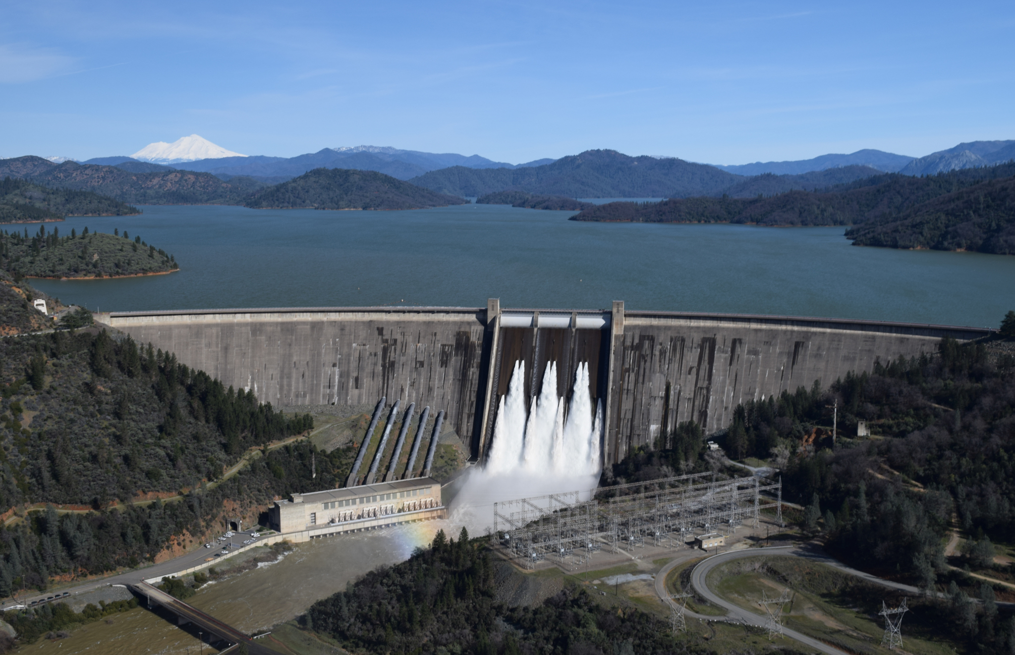 Shasta Dam releases water through its spillway as Lake Shasta rises following recent storms, with Mount Shasta visible in the distance.